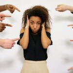 Stressed woman with hands on ears surrounded by pointing fingers, illustrating pressure.
